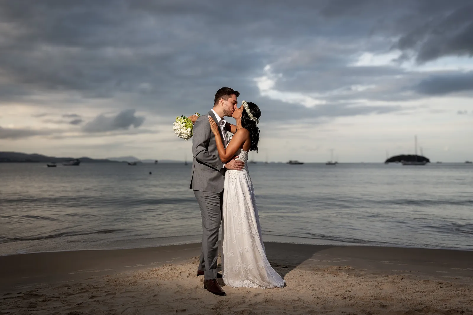 Casal de noivos se beijando na praia sob céu dramático, foto editada